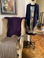 Full view of clothing lot including sleeveless Joseph Ribkoff dress on mannequin with navy jacket, purple cardigan and skirt on armchair with beige dress or top below.