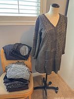 Gray long shirt with embroidered geometric pattern displayed on mannequin with several folded shirts on chair nearby