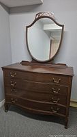 Front angled view of vintage mahogany 4-drawer dresser with attached mirror showing brass handles and curved front. Surface wear visible.