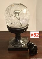 Photo of the snow globe on black pedestal and the small black glass elephant figurine together on a wooden surface, showing scale and shape.
