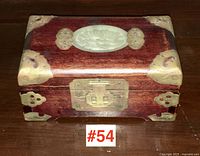 Front view of a small wooden Asian jewelry box with brass corner accents, brass latch, and oval carved jade inlay on lid.