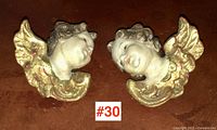 Pair of small Italian putti cherub head figurines shown together on a surface, both having gold-painted wings and painted faces.