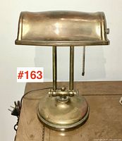 Front view of vintage brass plated desk lamp showing rounded rectangular shade, dual supporting rods, round base, and pull chain switch.