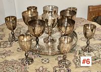 Ten silver-plated wine goblets arranged on a silver-plated tray on a patterned tablecloth.