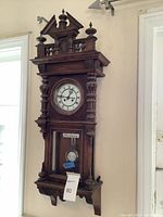 Front view of carved wooden wall clock showing pediment, dial, and lower pendulum window.