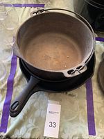 Photo showing a round cast iron pot with a bail handle sitting on top of a larger cast iron skillet, all on a patterned tablecloth.