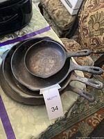 Photo showing all four cast iron skillets stacked inside each other, placed on a table with patterned chairs and carpet in background. Shows the varying sizes and well-used condition.