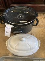 Black Crockpot with glass lid and electrical cord, resting next to large white ceramic serving dish with lid.