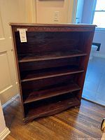 Full view of the dark wood book case showing four shelves and carved top edge, placed indoors against a wall.