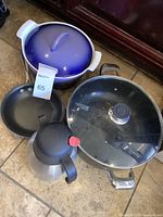 Photo showing blue covered pot, black frying pan, large pot with glass lid, and a small pouring carafe on tile floor