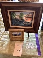 Two framed art pieces on table with size cards, larger painting of a riverside buildings scene and smaller landscape painting