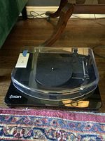 Black Ion record player on a wooden floor with carpet underneath, showing the entire player with closed clear plastic dust cover.