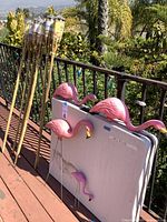 Full view of four tiki torches and four pink plastic flamingo decorations leaning against a railing outdoors.
