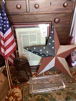 Lot photo showing the framed document, wooden eagle statue, metal patriotic star, American flags, and Scottish terriers license plate cover on a carpet in front of a wooden chest.