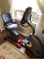 Full view of black Schwinn 270 recumbent exercise bike showing seat with manual and dumbbells on frame, positioned by window.