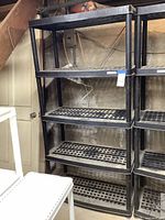 Full view of the tall black plastic storage rack showing four grid-patterned shelves and thick vertical supports. Located in a basement area with concrete walls and visible utility wires.