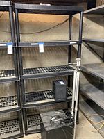 Photo of the tall black plastic storage rack showing all four slotted shelves and cylindrical legs, with other racks adjacent.