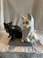Front-left angle view of black and white glossy porcelain cat figurines seated on a grey display base against a neutral fabric background.