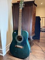 Front view of teal Hohner acoustic guitar showing body, neck, and headstock above wooden floor and near wooden cupboard.