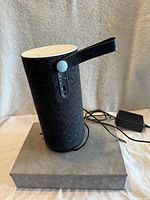 Front side view of the black felt cylindrical speaker with white top, control panel, black leather handle, and power charger visible on grey display base