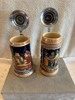 Photo of two German beer steins showing inside metal lids in open position, front side with hand-painted drinking scene and inscription 'NACH DANNI'
