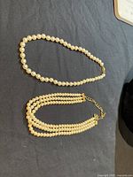 Both pearl necklaces shown against a black background. The single strand necklace is a uniform size of pearls with a simple gold-tone clasp. The other is a triple strand with smaller pearls and a gold-tone multi-strand clasp with extender.