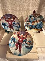 Photo of 3 round Stewart Sherwood hockey collector plates showing detailed hockey scenes and player celebration, placed on original boxes on a beige fabric background.