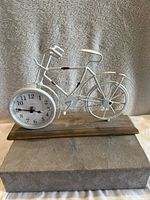 Front view of rustic metal bicycle mantel clock with clock face as front wheel, mounted on wooden base, displayed on grey textured fabric.