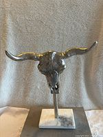 Front view of silver-toned metal bull/cow skull sculpture with horns on white marble-like stone base, displayed against a neutral fabric background.