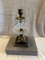 Front angled view of the ornate brass candle holder resting on a gray box, showing the detailed base, crystal drip tray and hanging prisms clearly.