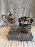 Glass coffee carafe with silver plated handle and lid, sitting next to silver plated candle warmer stand on gray base.