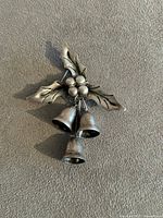 Front view of a vintage sterling silver brooch with detailed holly leaves, berries, and three dangling bells on a textured neutral background.
