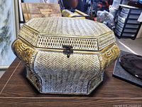 Front and side view of an octagonal wicker basket chest with hinged lid and sculpted handles, showing the locking clasp detail.