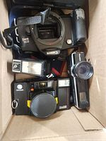 Box containing a Canon EOS film camera body, Minolta camera with attached flash, another vintage camera, and camera flash units.