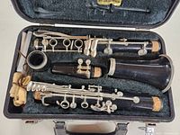 Clarinet disassembled inside black case with fabric lining; visible cork joints and silver keys; mouthpiece shows one crack.
