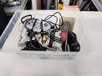 Top view of a plastic bin containing multiple wired video game controllers of different styles including a Nintendo 64 controller and classic joystick.