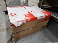 Shows the box containing the 10 reams of Staples copy paper with red and white packaging, indicating the brand and copy paper type.