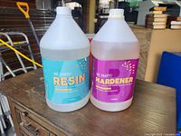 Photo of the front of two 1-gallon containers, one labeled Dr. Crafty Resin and the other Dr. Crafty Hardener, placed on a wooden surface with some background items.