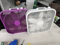 Two box fans on table, showing white and purple color, both with handles and grills.