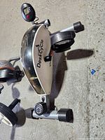 Desk cycle exerciser showing side view, compact black and white design with pedals and adjustable resistance.