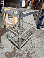 Full view of small metal wire rack shelf showing all three shelves and structure, placed indoors on concrete floor with some items in background.
