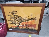 Full view of the framed stitch art depicting a tree at sunset with birds flying and hills in the background.