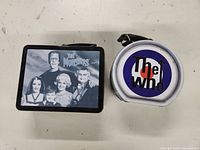 Front view of two tin lunchboxes side by side. Left: The Munsters rectangular box showing characters. Right: The Who round lunchbox with logo.