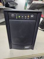 Front panel view of Tripp Lite UPS showing power button, status LEDs, and mute button, with the main body in black plastic casing.