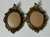 Front view of two ornate vintage brass oval photo frames with intricate decorative detailing around oval window and side handles, cardboard inserts inside frames.
