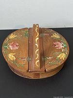 Front view of oval wooden box showing folk art hand-painted floral decoration and central hinged section with wavy painted stripes.
