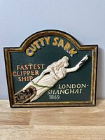 Front view of vintage wooden Cutty Sark plaque with carved relief of a woman and yellow lettering on green background.