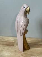 Hand carved wooden parrot figurine painted light pink with golden beak and perch, showing side view.