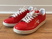 Pair of red vinyl Adidas Stan Smith sneakers with white heart design on the sides. White laces and midsoles. Shoes show minor wear and scuffs.