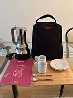 All components laid out on table: espresso maker on base with cord, Alessi carry case, two cups, two saucers, two spoons, manual